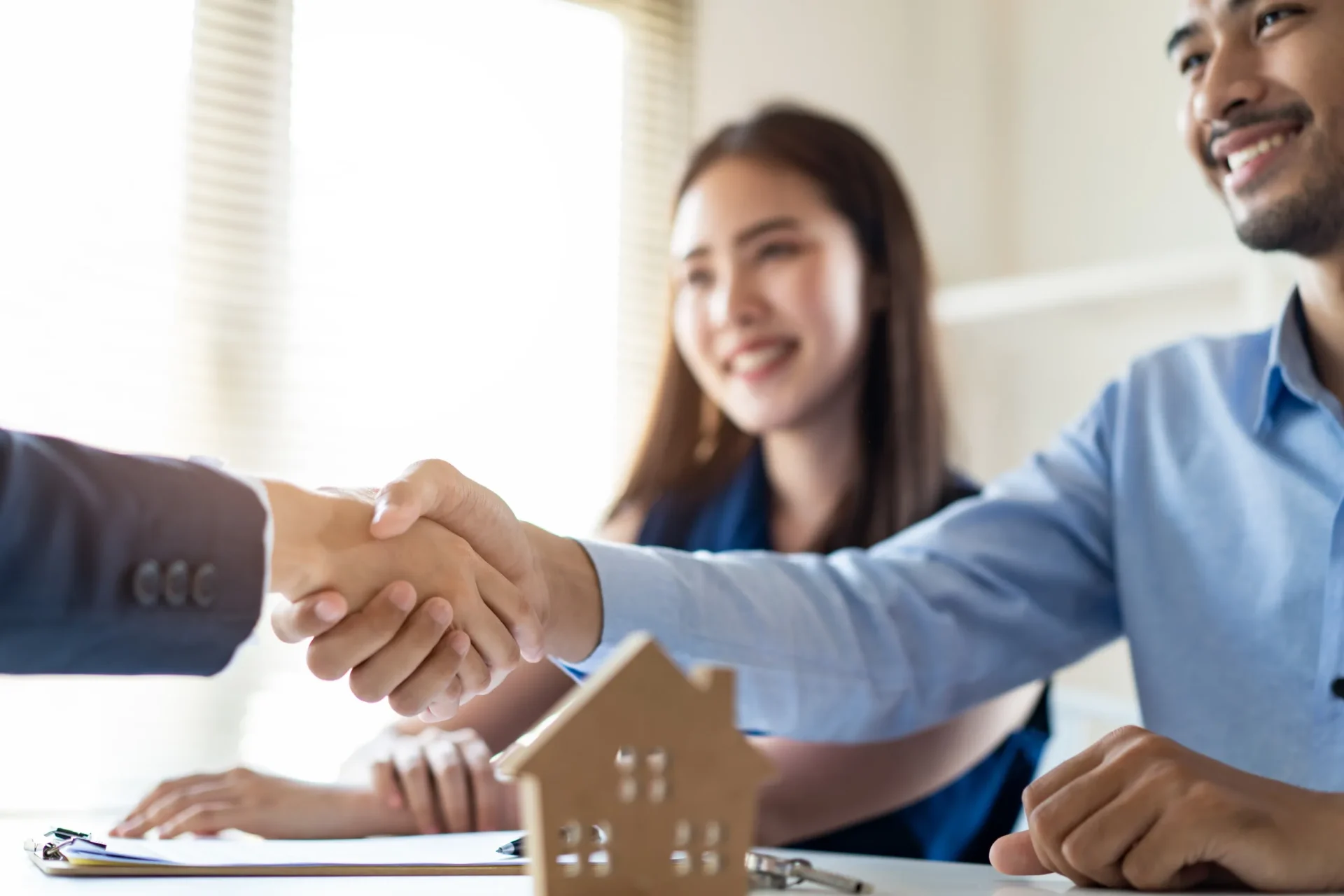 Young Asian couple making contract with house sale agency. Man shaking hand with female agent sitting opposite and his wife sitting next to him with smile looking at house broker. Real estate concept.